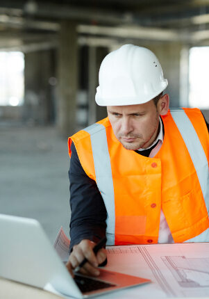 Portrait of development supervisor wearing protective vest and helmet over formal suit  proofing blueprints using laptop computer inside unfinished building